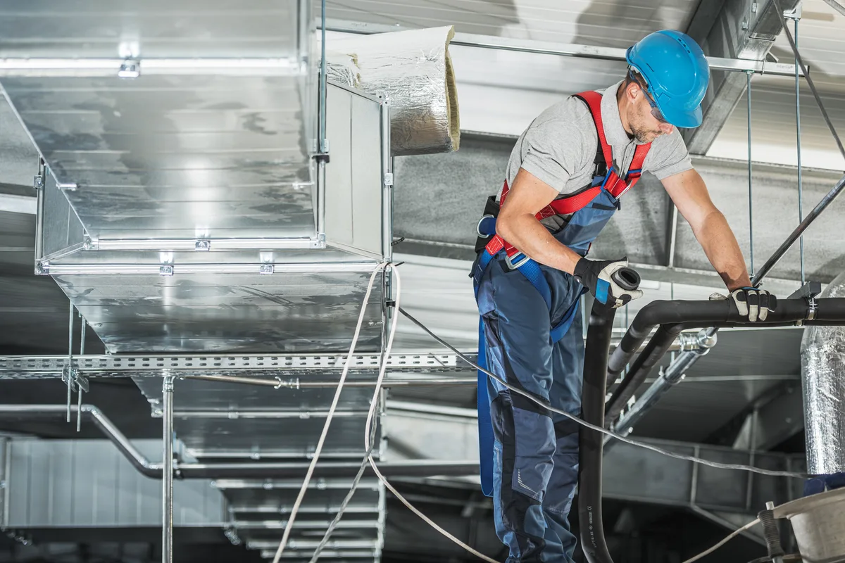 An HVAC technician in a hard hat handling flexible duct beneath a run of sheet-metal ductwork — HVAC contractor insurance in Alabama.