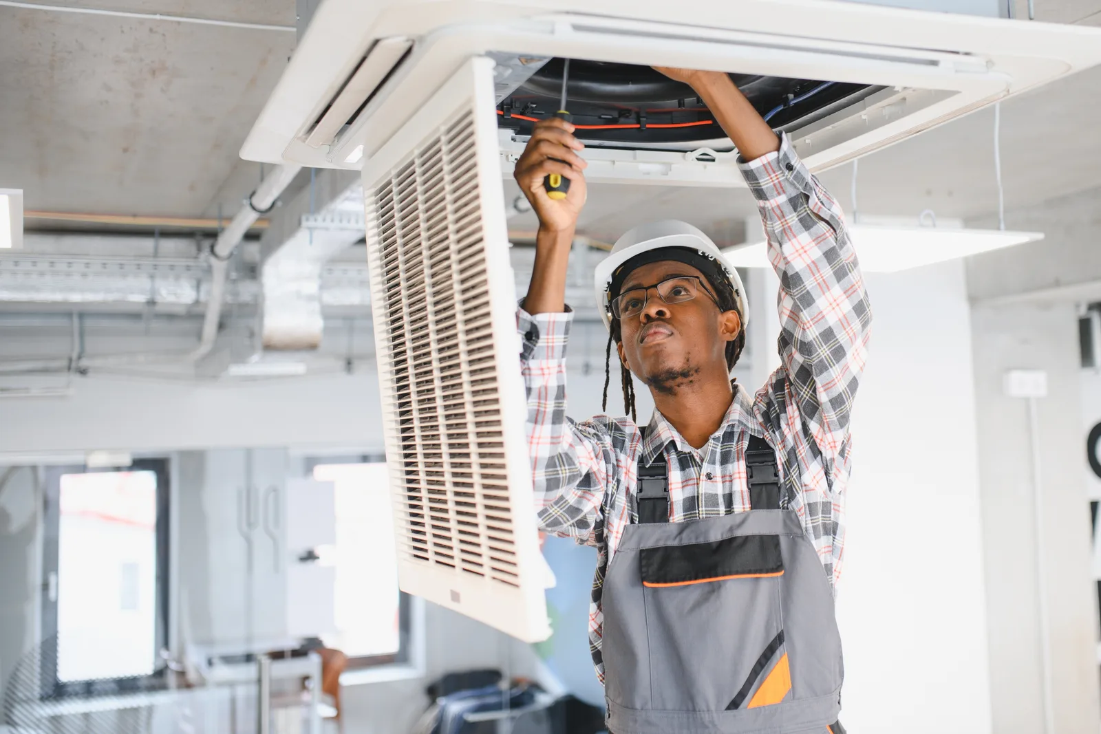 An HVAC technician in a hard hat servicing an open ceiling-cassette unit from below — HVAC contractor insurance in Illinois.