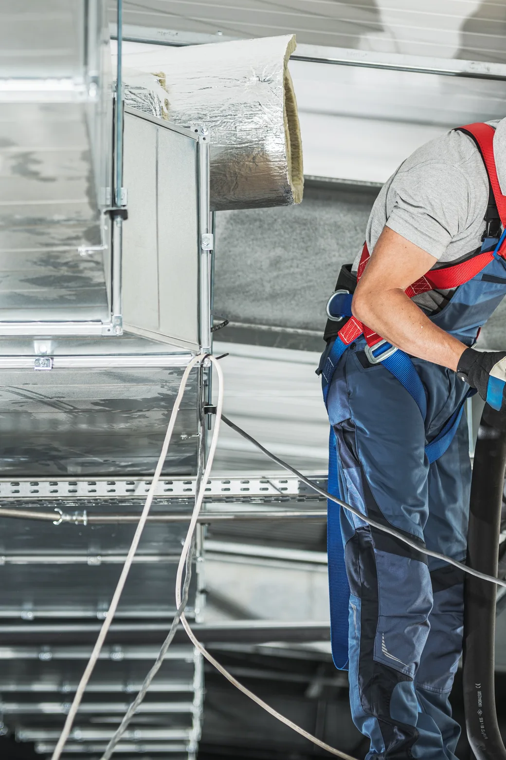 An HVAC technician in a hard hat working beside overhead sheet-metal ductwork in a mechanical space — HVAC contractor insurance in Texas.