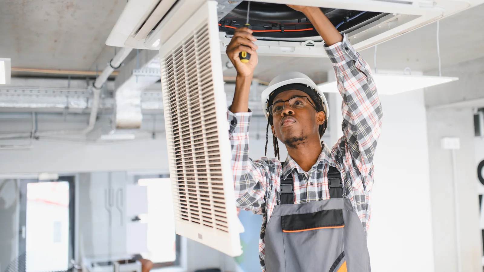 An HVAC technician reaching into the open grille of a ceiling-cassette unit in a commercial interior