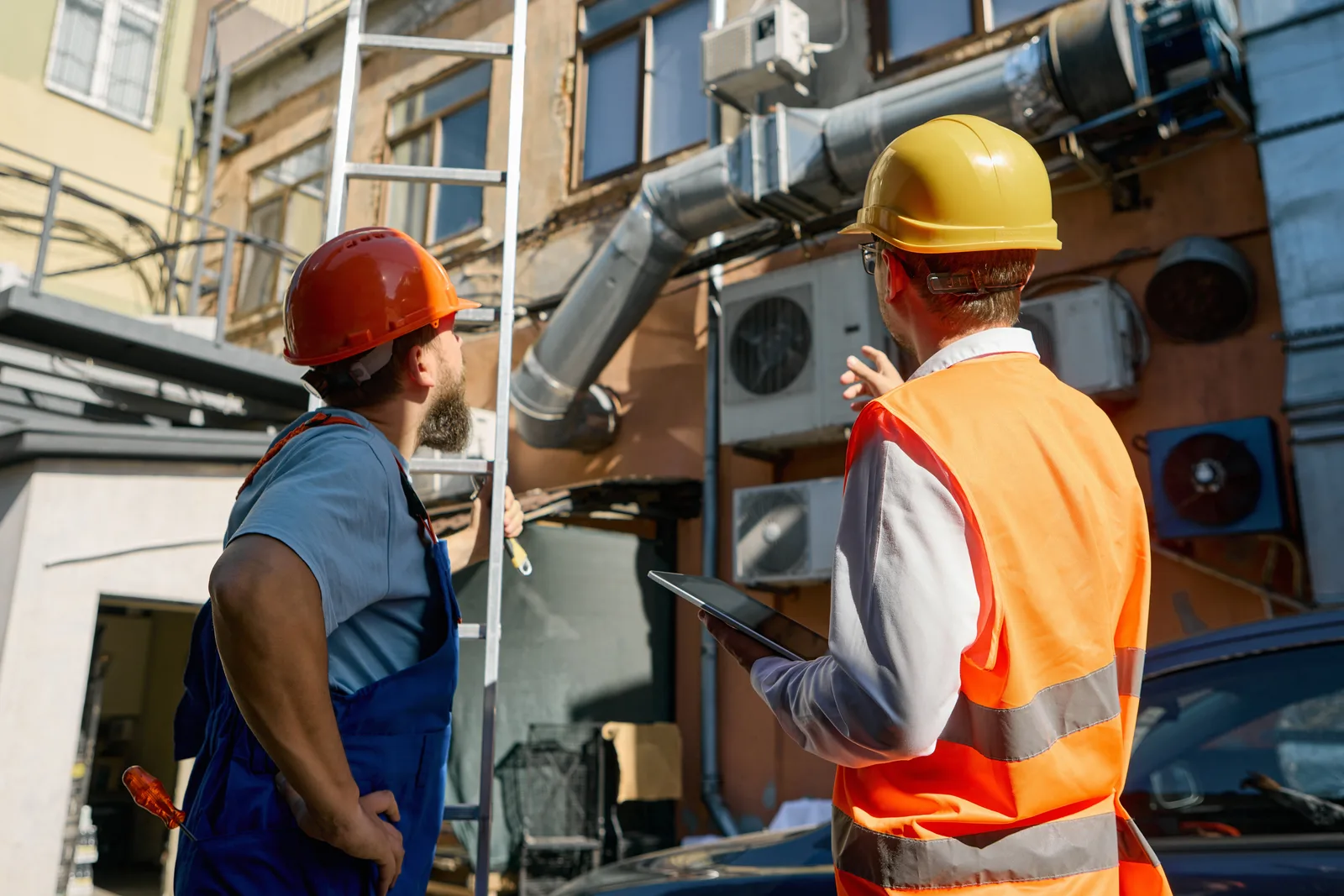 Two workers in hard hats reviewing exterior ductwork and wall-mounted condensing units — HVAC contractor insurance in Nebraska.