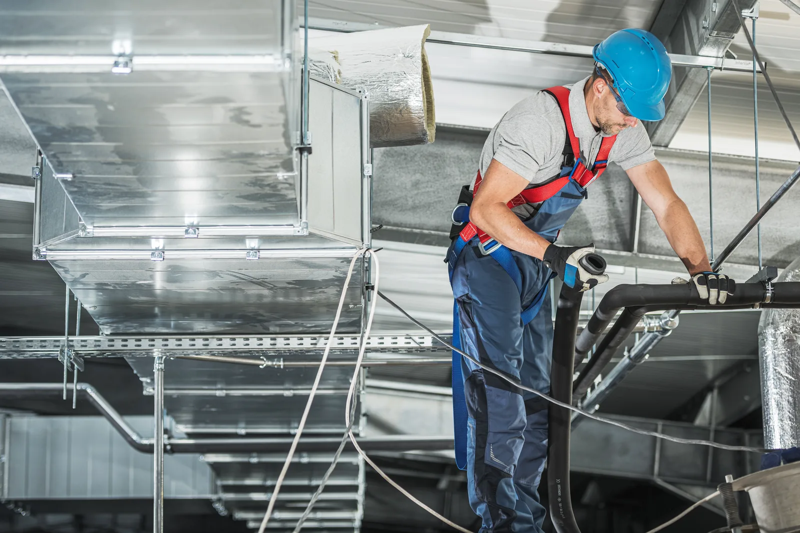 An HVAC technician in a hard hat pulling flexible duct beside overhead sheet-metal trunk lines — HVAC contractor insurance in Georgia.